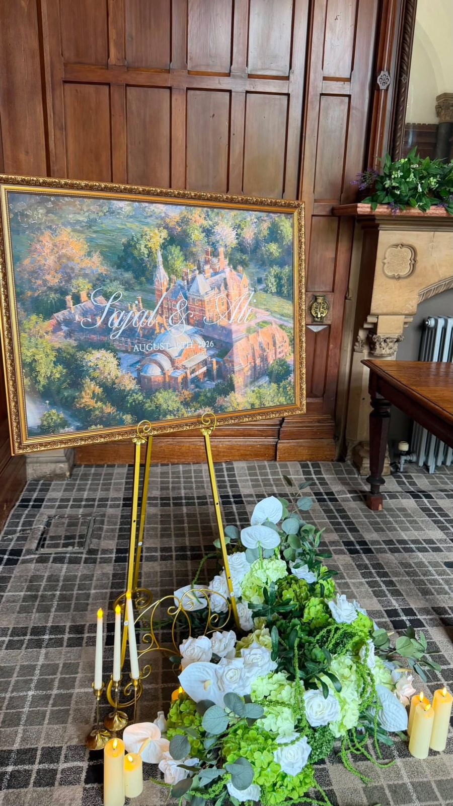 Hand-painted venue portrait on a gilded easel framed by white roses and candles